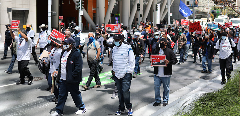 Masked people marching outside. Some have signs: "UNLOCK THE MONEY," "FREE OUR FUNDING," "AUSTERITY IS RACIST!"