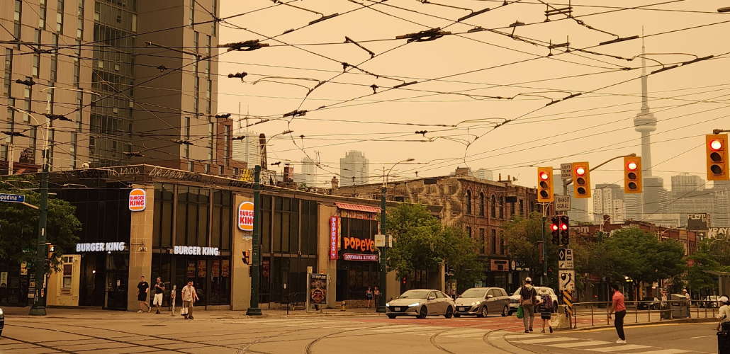View of a city intersection. The sky, crisscrossed with power lines, is hazy grey and eerily tinted orange. Some people are out walking anyway.