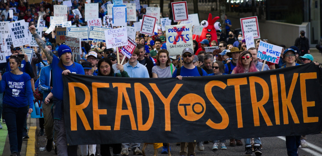 Big crowd marches behind a giant black banner hand-painted in orange "Ready to Strike." Many people hold handmade signs on sticks. A person near the front with long pink hair and reflecting sunglasses looks defiantly into the camera. A person in a blue beanie punches a tattooed arm into the air, raising a fist. Farther back, glimpse someone in a red inflatable lobster suit.