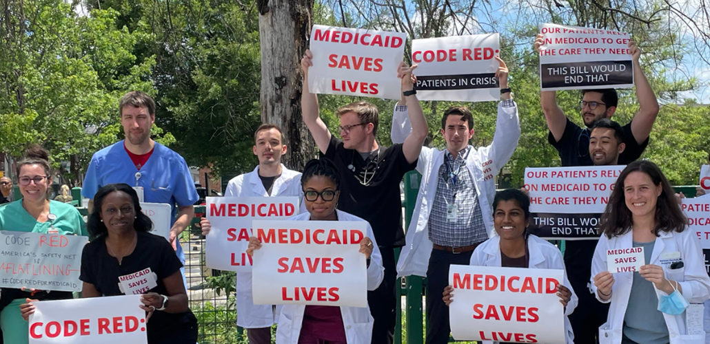 A diverse group of people, mostly in white lab coats, stand outside holding printed signs that say, in red on white, "Medicaid saves lives." Other signs say "Code red: patients in crisis" and "Our patients rely on Medicaid to get the care they need. This bill would end that."