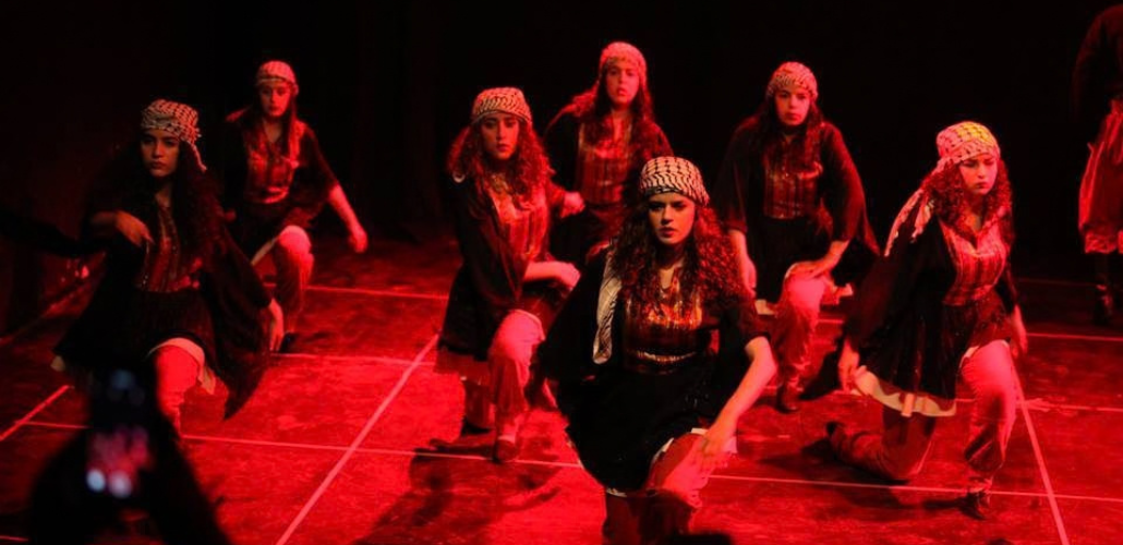 Seven women in long skirts and kerchiefs kneel, their arms in motion, apparently mid-dance. They are on a stage, bathed in red light.