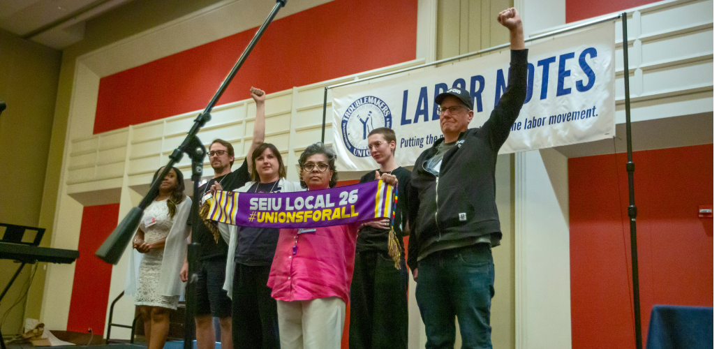 Six workers, of various races and genders, stand together on a ballroom stage with a big "Labor Notes" banner hanging behind them. Three are raising fists in the air, and one holds out a purple "SEIU Local 26, #UnionsForAll" scarf.
