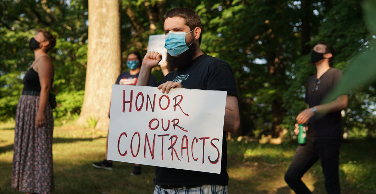 Masked person holding sign "Honor Our Contracts" in front of trees, other masked demonstrators