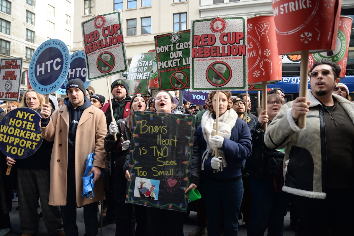 A tight-packed group stands with signs and giant Starbucks cups.