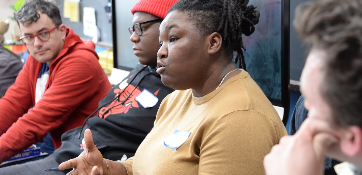 A Black woman in a tan shirt speaks while three men, white and Black, look on.