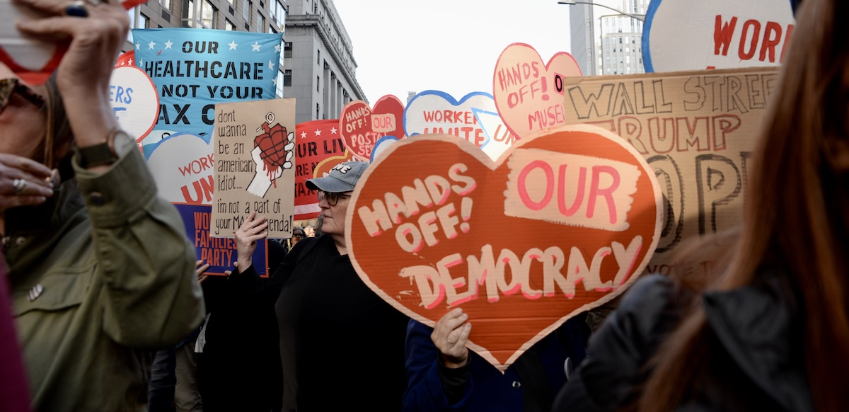A heart-shaped sign saying “Hands Off Our Democracy” is carried in a crowd.