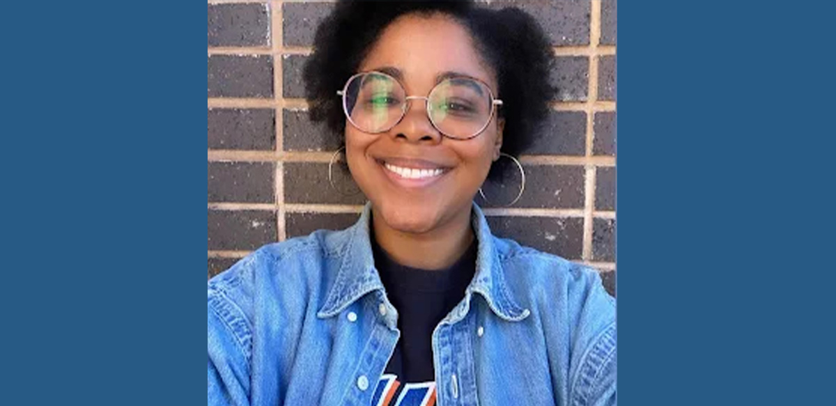 A smiling Black woman in round glasses and a jean jacket is shown