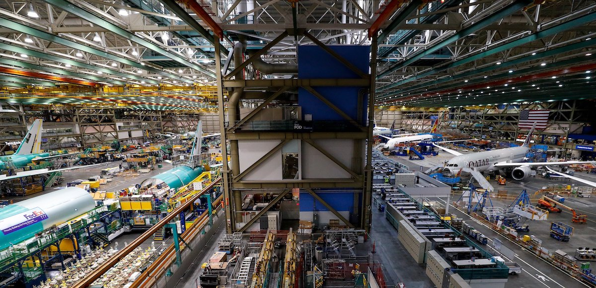 An aircraft factory floor is viewed from above, with planes on either side and high rafters above.