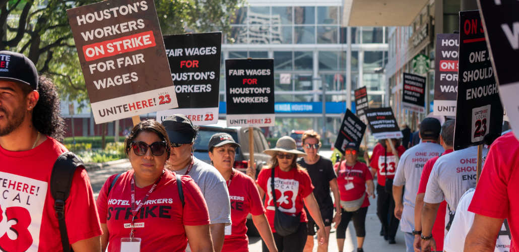 People in red T-shirts picket in the sunlight on a downtown sidewalk. Shirts say "Hotel workers strike" and "UNITE HERE Local 23." Picket signs say "Houston's workers on strike for fair wages." Another slogan partially visible on some signs is "One job should be enough."