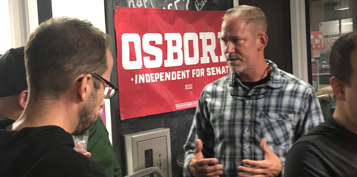 A man in a checkered shirt talks to two people with their backs to the camera in front of a red “Dan Osborne Independent for Senate” banner.