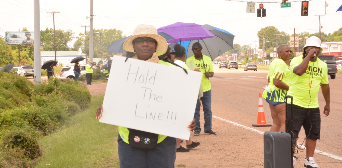 A woman holds a sign reading Hold the Line while others stand on the picket line.