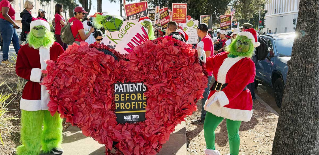 Two people in green Grinch suits hold a giant paper-mache heart that contains a "patients before profits" sign. Behind them an NUHW Kaiser picket line crowd is visible.