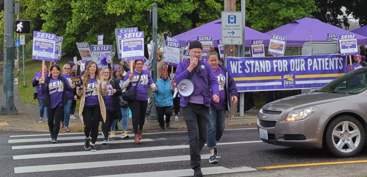 Workers in purple SEIU Local 49 T-shirts march across a street. Some carry picket signs that say SEIU and some other words too small to read. A large purple banner says "We stand for our patients." The person in front is talking into a bullhorn and others appear to be chanting. 