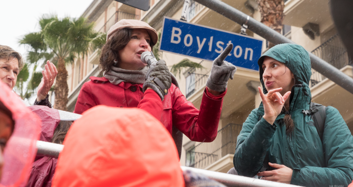 A woman speaks at a strike rally while another woman interprets in sign language. Behind them is a palm tree, an apartment buidling or hotel, and a street sign "Boylston"