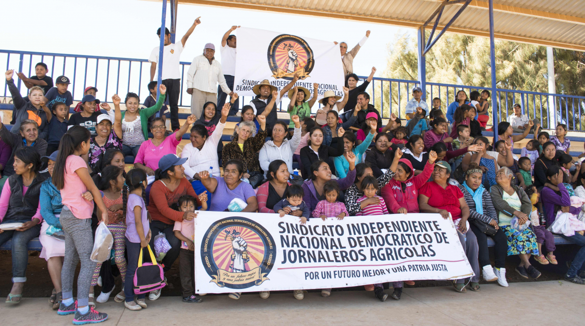 A group of Mexican women farmworkers sit in several rows behind the SINDJA banner.