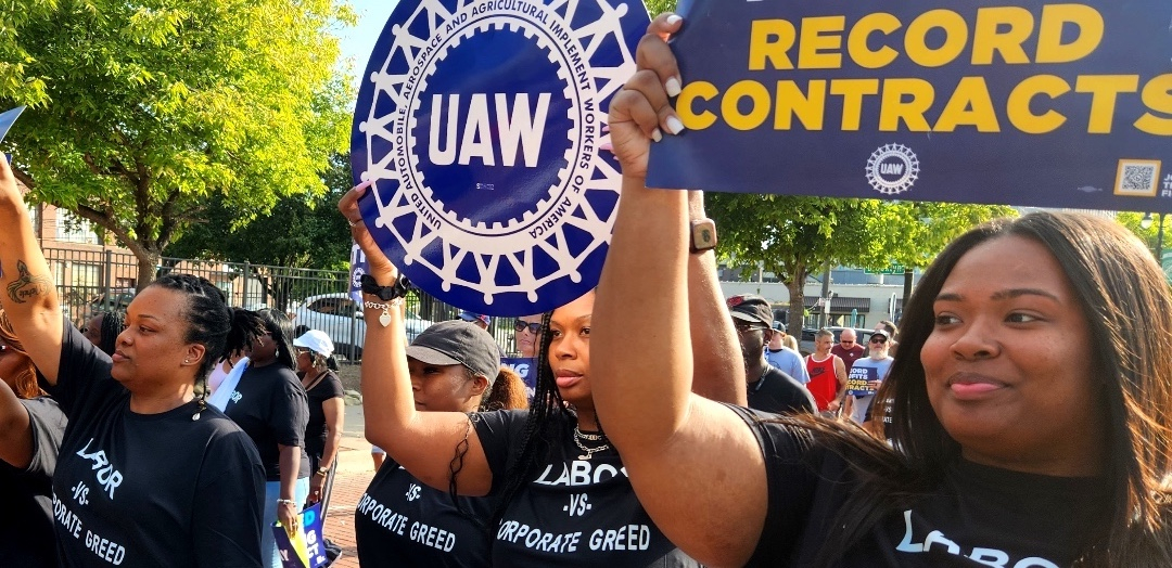 Four Black women march with t-shirts that say “Labor vs. Corporate Greed.” They’re holding UAW signs.