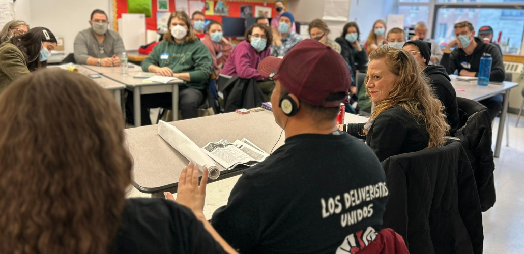 People sit in a classroom. Most are looking attentively at a person in the foreground who must be speaking, but has his back to us. He is wearing an interpretation headset and the back of his T-shirt says "Los Deliveristas Unidos," Spanish for united delivery workers.