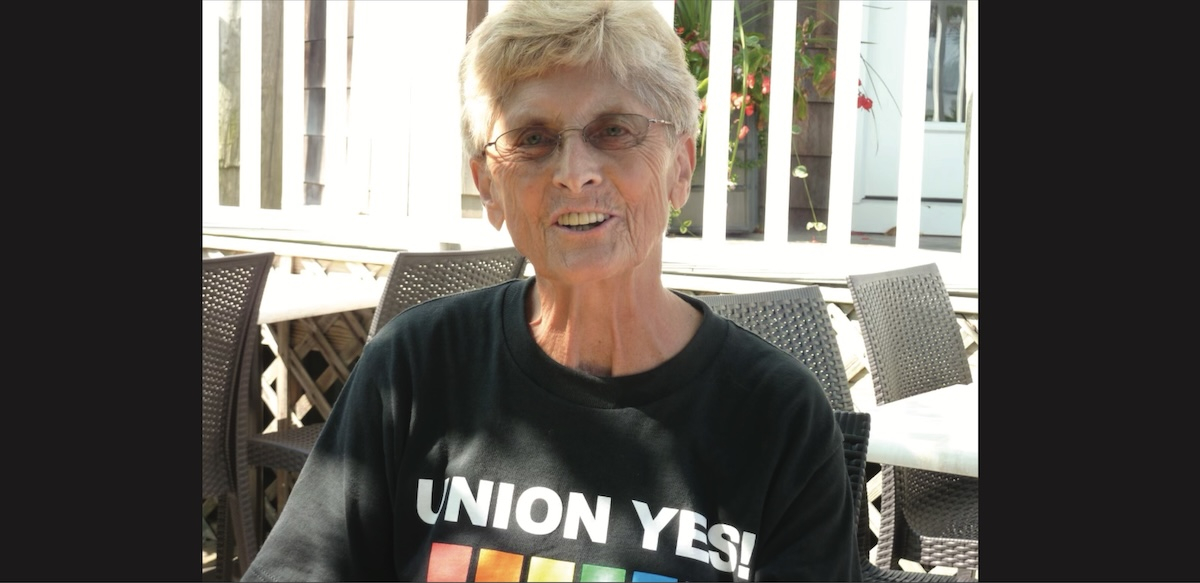 A smiling woman is pictured with short blond hair and a black Union Yes t-shirt with a rainbow on it.