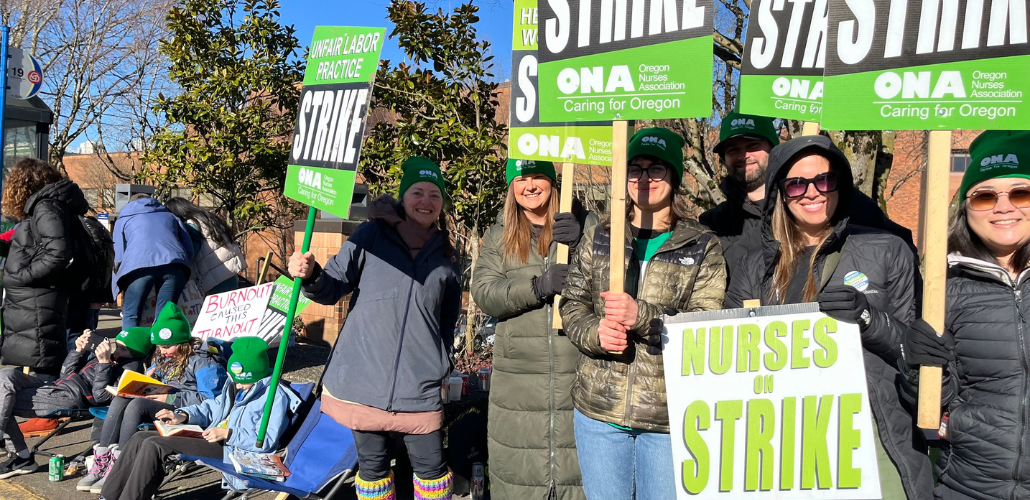 Women stand on a sunny day, wearing big jackets, holding green printed ONA picket signs that say "Unfair Labor Practice Strike" and "Nurses on Strike." A handmade sign visible in the background says "Burnout caused this turnout."