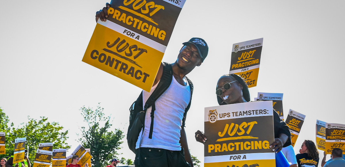 A young slender Black man in a baseball cap smiles down at the camera with a sign saying “Just Practicing for a Just Contract.” To the left of him is a Black woman in sunglasses with a whistle in her mouth carrying the same sign..