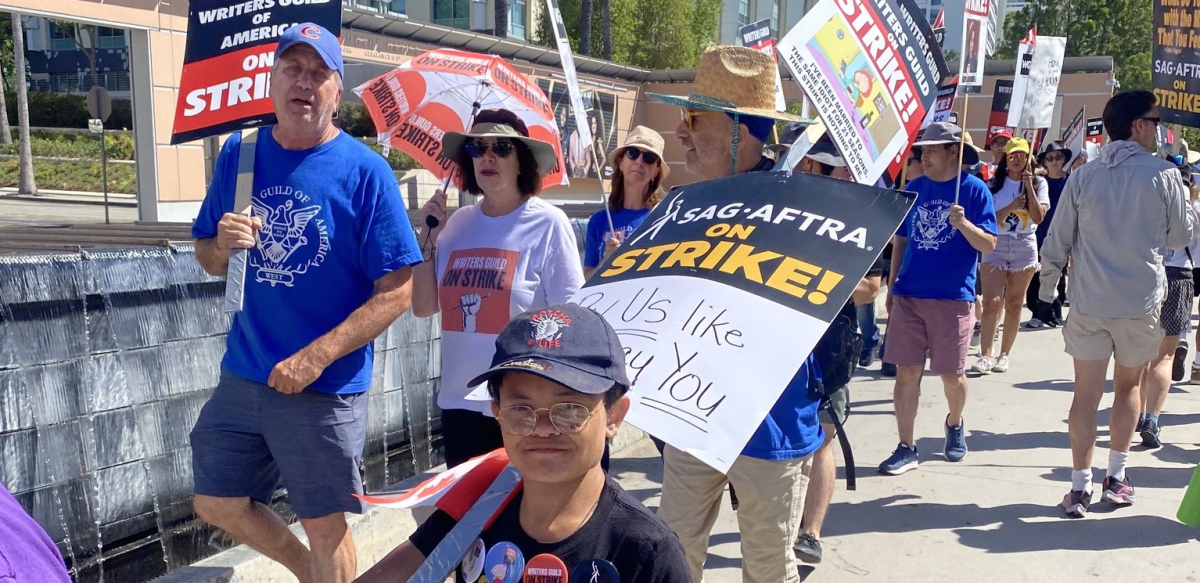 Actor Sam Humphrey and several others march towards the camera with ‘SAG AFTRA on strike’ and ‘Writers Guild of America On Strike’