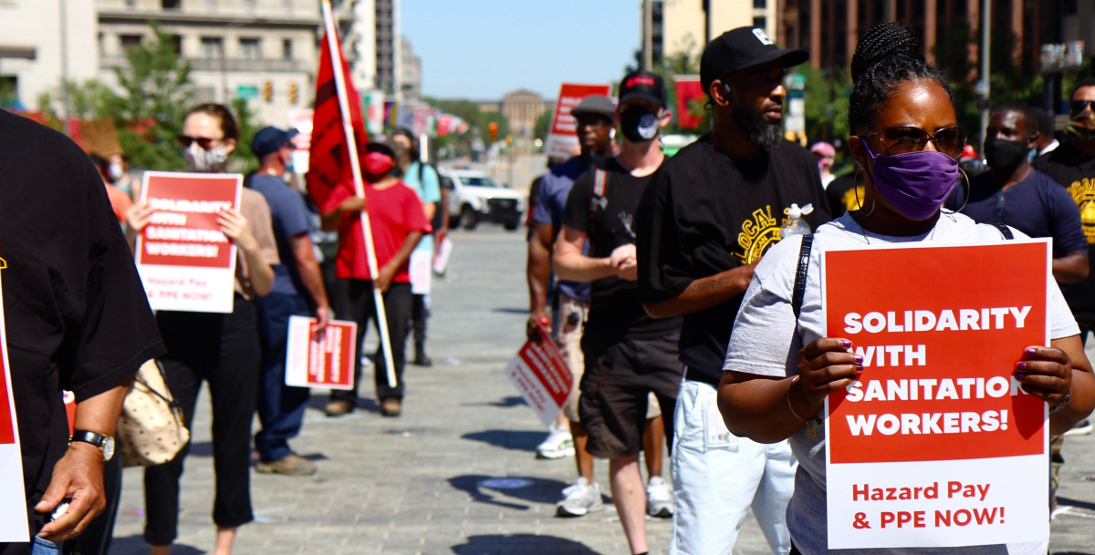 Masked people stand outside holding red printed signs. Visible sign in foreground says: "Solidarity with Sanitation Workers! Hazard Pay and PPE Now!"