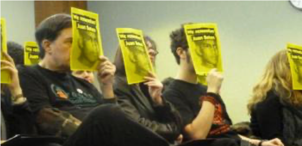 Protesters sitting in a row in a meeting hold up yellow posters with a picture of slain worker Juan Baten.