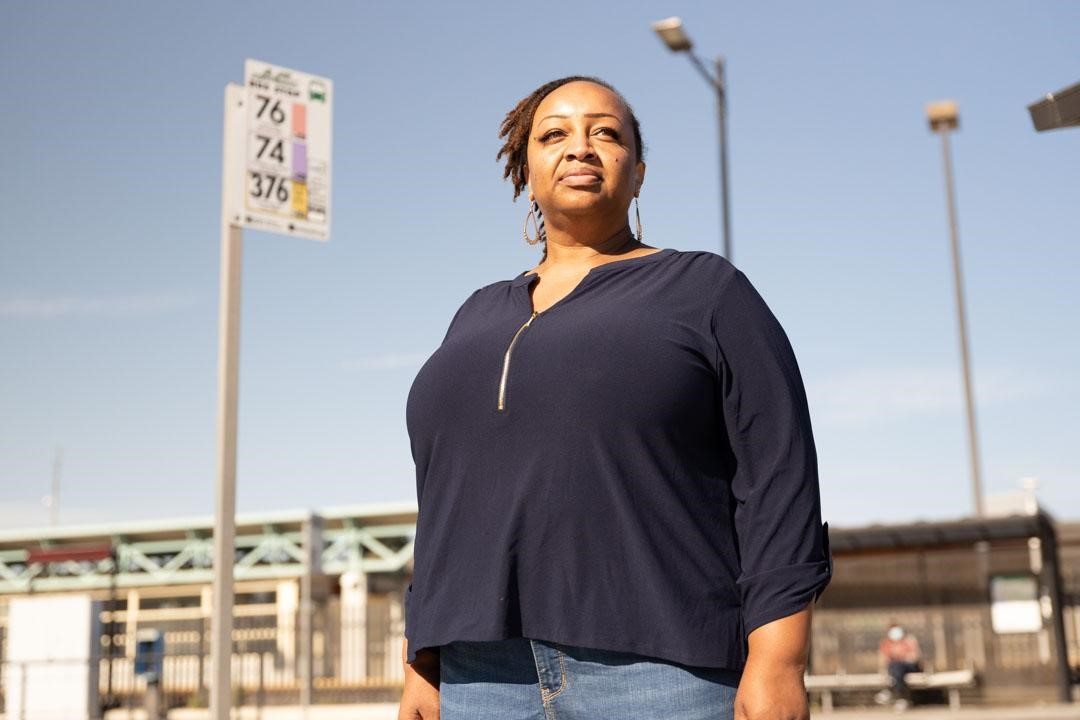 Black woman looks into the distance with a serious, determined expression. Behind her is a bus stop sign and a blue sky.