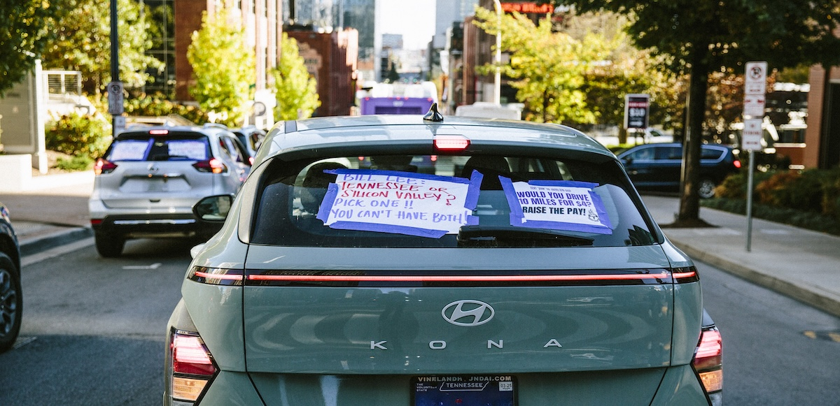 A car back window carries signs, including one saying, “Would you drive 10 miles for $4? Raise the Pay!”