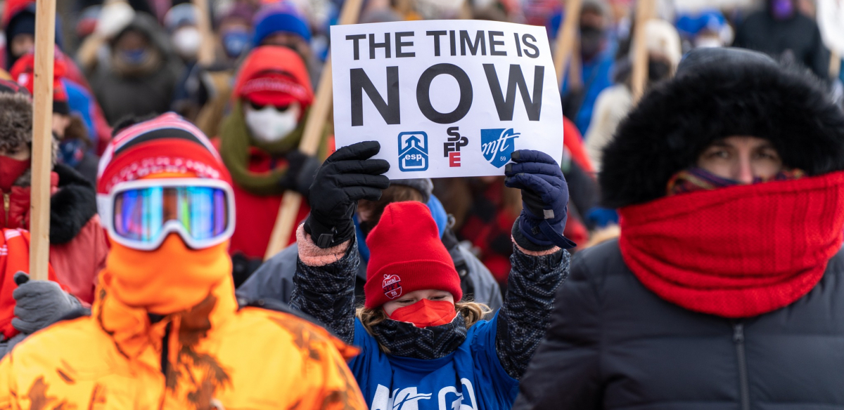 Marchers bundled in coats, one carries printed sign: "The time is now!"