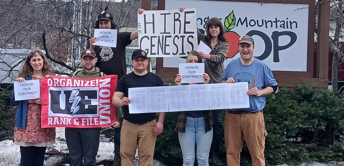 Eight people outside face the camera with signs, including “Hire Genesis” and a UE banner.