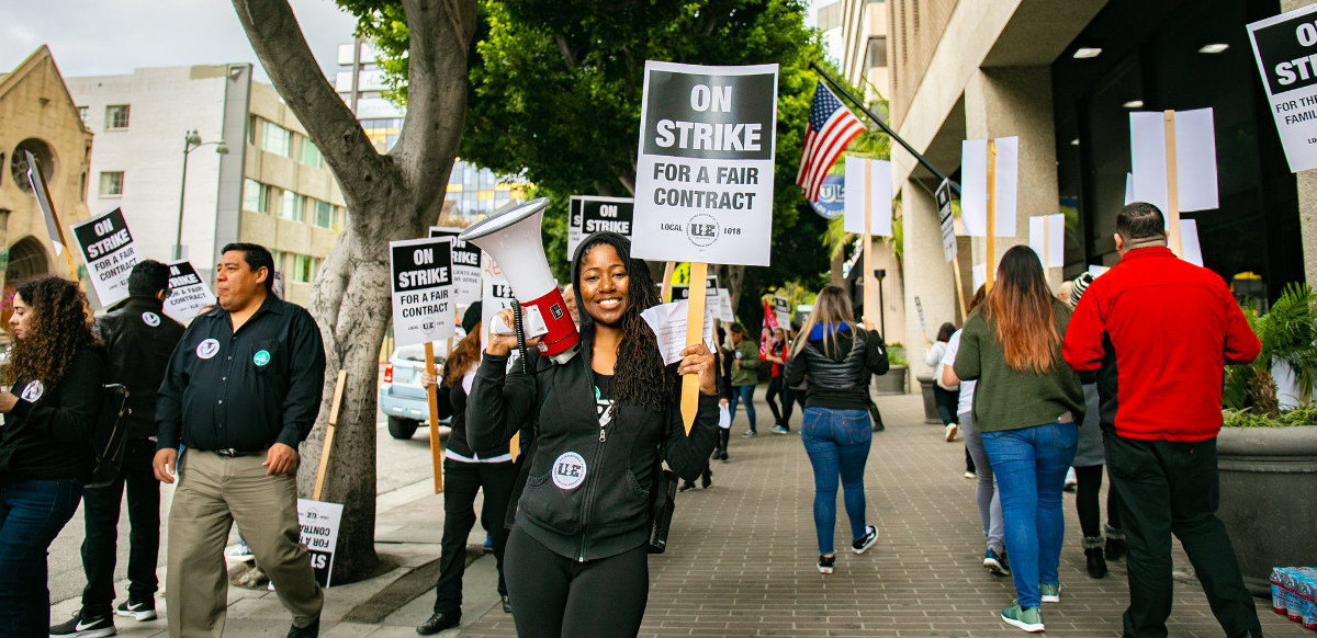 A woman holds a bullhorn and a picket sign that says "On Strike for a Fair Contract," surrounded by dozens of picketers holding similar signs and marching in a circle on the sidewalk.