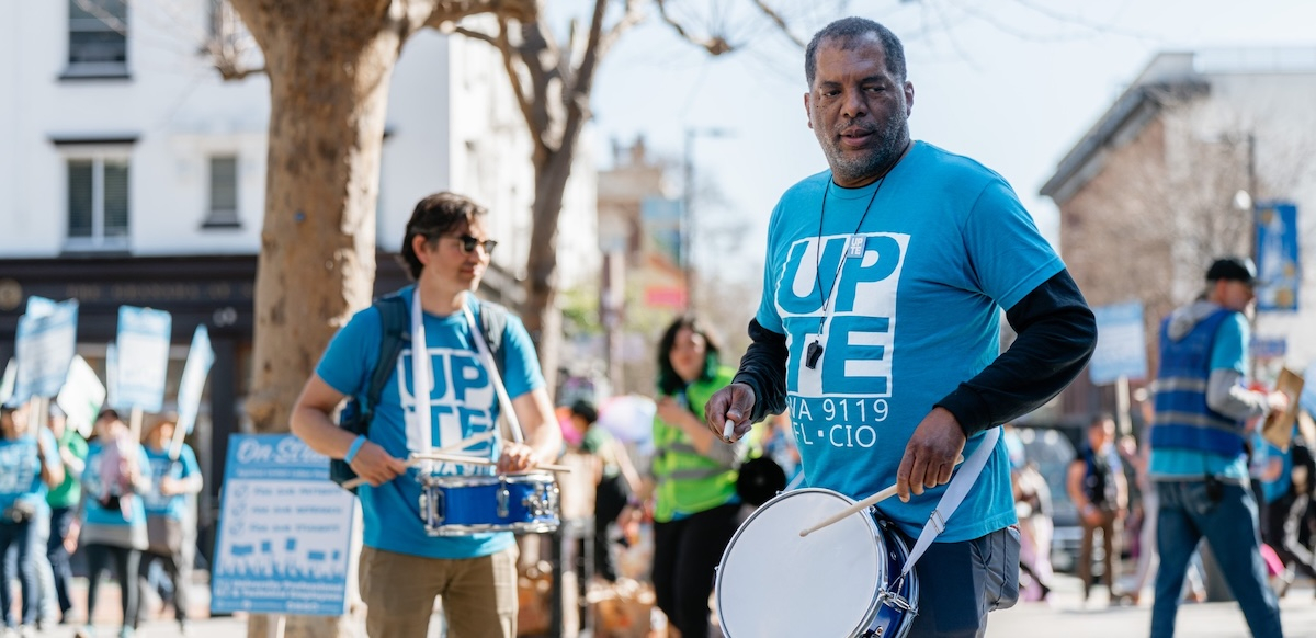 Two men in blue shirts that say “UPTE” play snare drums while others march in the background.