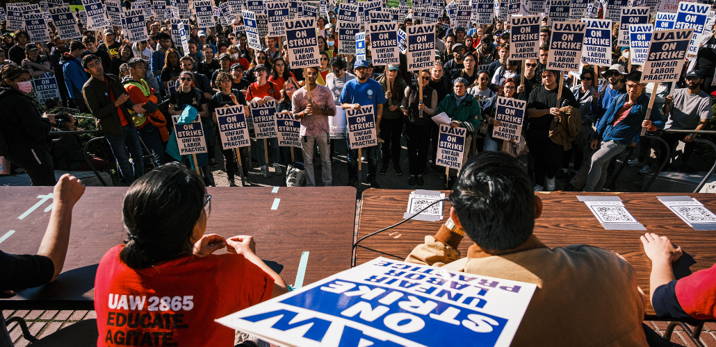 In the foreground we see the backs of a few strikers in red UAW 2865 T-shirts, one holding a strike sign, sitting at a table. Facing them is a huge crowd of strikers, standing, with picket signs, outdoors. The signs say "UAW on strike, unfair labor practice."