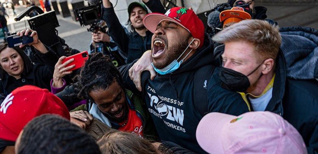 Tight crowd of people outdoors embracing and yelling in celebration. Most visible person in center, face upturned, wears a red baseball cap and a T-shirt that says The Congress of Essential Workers. Behind, many people hold up cameras capturing the moment.