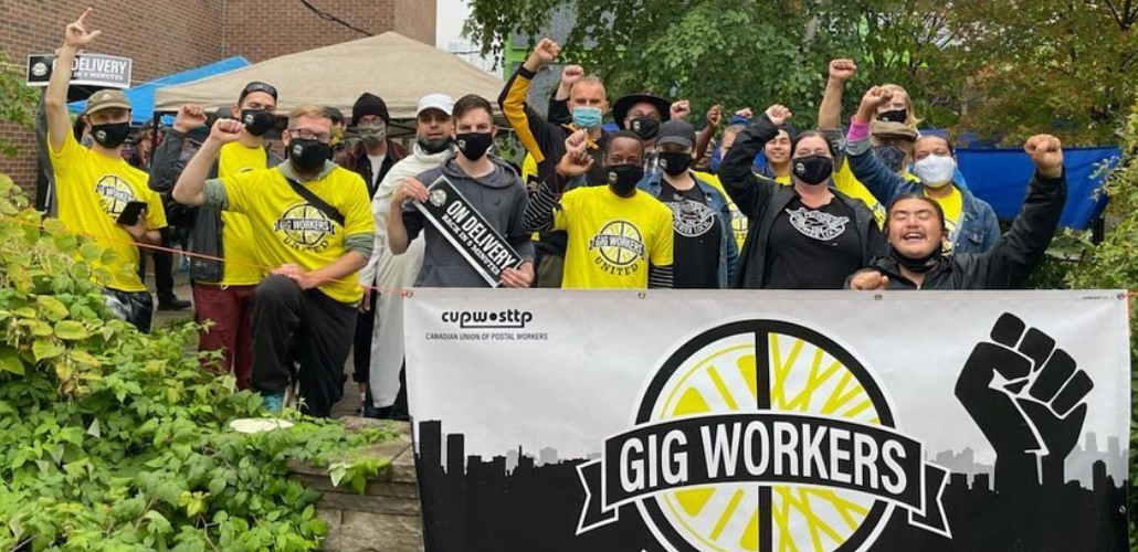 Young people of various races, fists in air, all masked. Some wear yellow "Gig Worker United" shirts and those in front hold a big banner with the group's logo and the initials of the Canadian postal union.