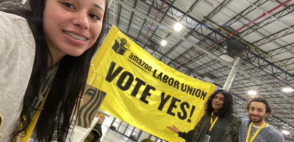Inside a warehouse three young people smile as they unveil a bright yellow banner that says "Amazon Labor Union, Vote Yes"