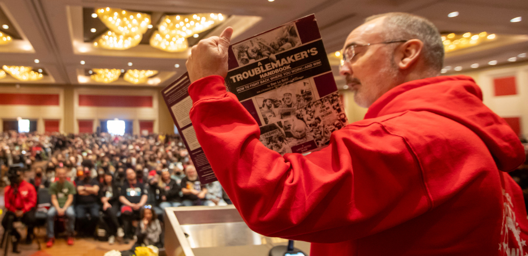 Seen from the side/back, Shawn Fain in a red hoodie holds up a battered purple copy of A Troublemaker's Handbook. Arrayed in front of him can be seen a huge seated crowd in the ballroom for the closing plenary.