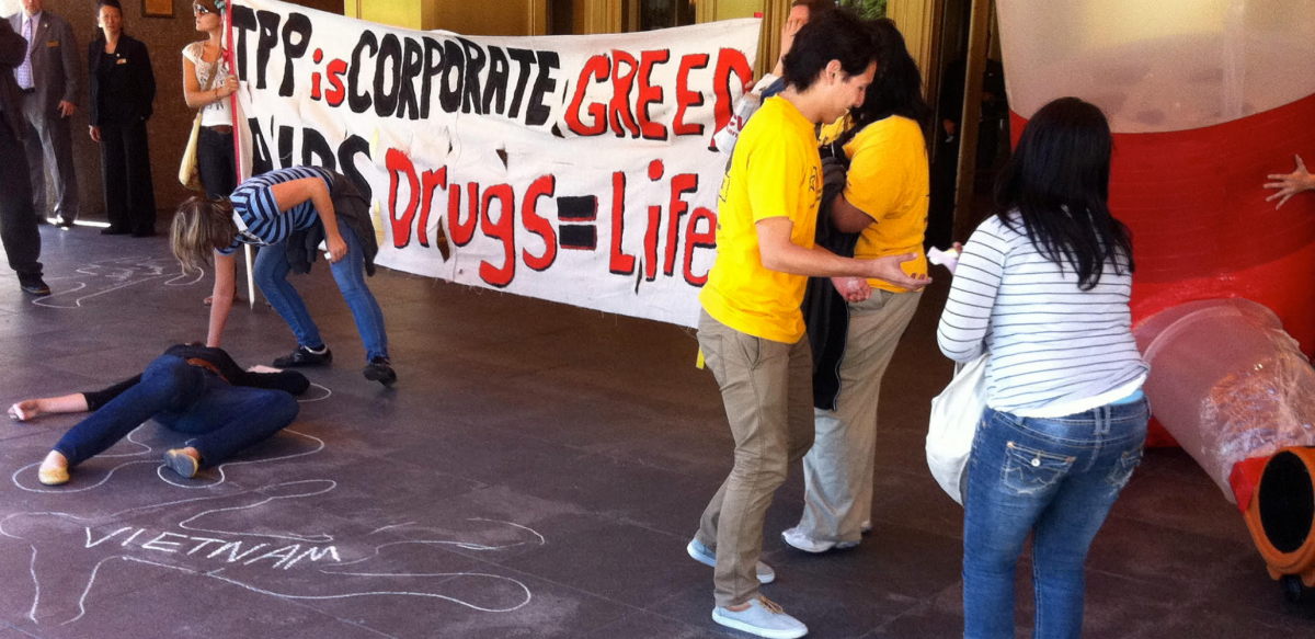 Activists draw chaik outlines on the ground in front of a hotel entrance; one outline is labeled "Vietnam." Others hold a banner: "TPP is corporate greed. AiDS drugs = life." 