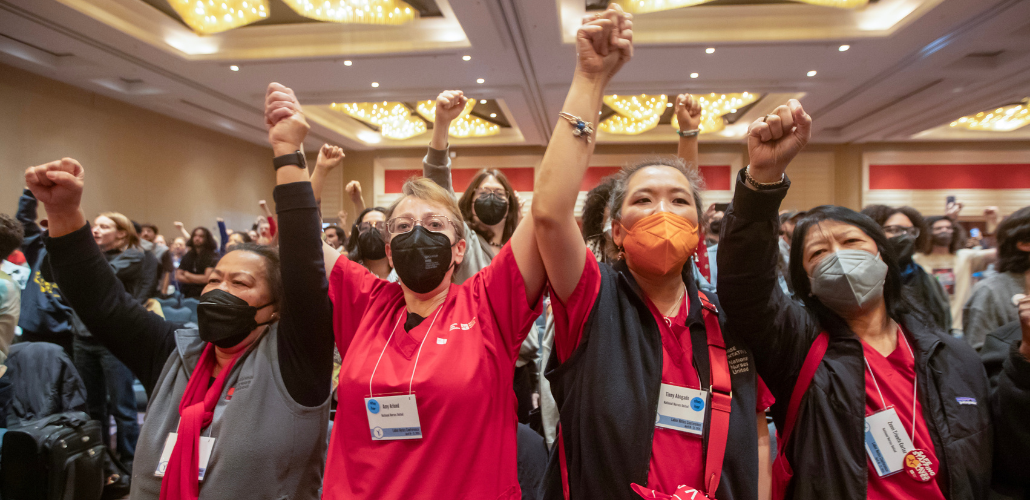 Four women in red scrubs, of various races, stand, all masked, raising their fists in the air, some clasping one another's hands. They are at the front of a large crowd in the ballroom.