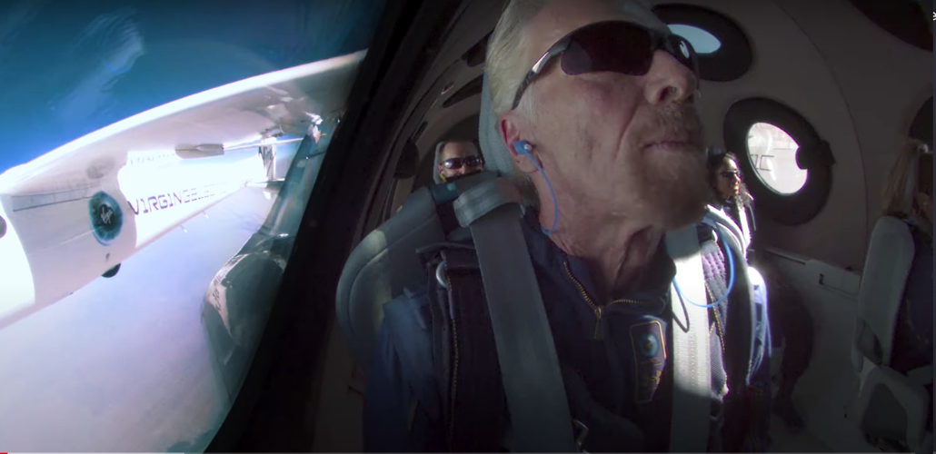 Screenshot of Richard Branson, wearing sunglasses and earphones, in a rocket. Two other crew members are visible behind him. On the left is a window through which you can see part of the outside of the rocket, labeled VIRGIN, and blue sky.