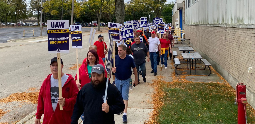 A dense line of picketers (at least 25 people are visible but it looks like there are more behind them) marches towards the camera on a leafy sidewalk alongside a factory. Many carry printed signs like "UAW on strike" and "Stand up for retirement security." Most visible faces are white but a few Black faces are also visible; there's a mix of women and men.