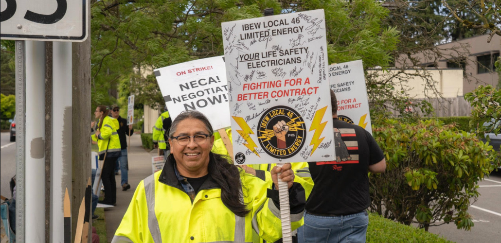 Picketers picket on a sidewalk. Person in foreground, wearing a safety-yellow jacket, smiles at the camera. This person has brown skin, glasses, hair in a ponytail, and holds a Local 46 picket sign printed with a limited energy of a fist holding bolts of lightning and the words "Your Life Safety Electricians, Fighting for a Better Contract." The sign appears to be autographed by many other strikers. Another printed sign visible in background says "On strike! NECA! Negotiate now!"