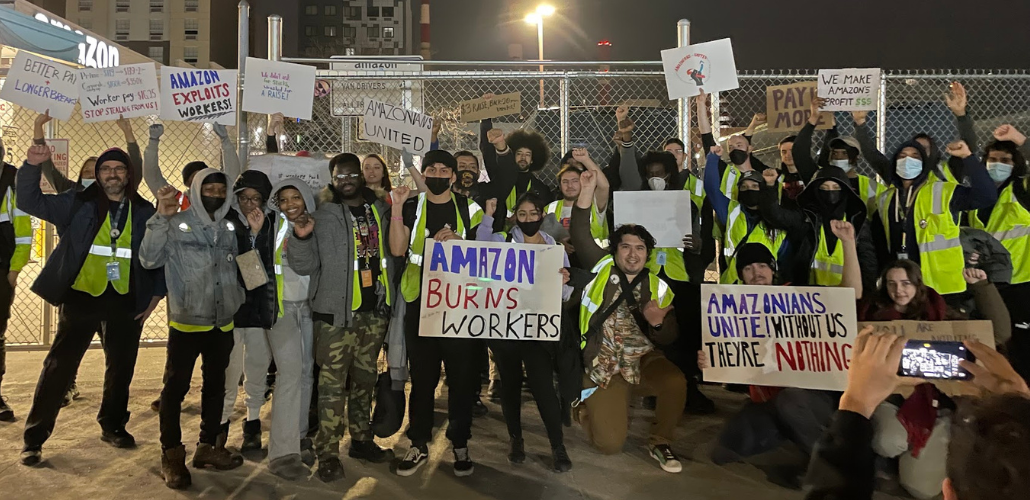 Crowd of excited owrkers stands outside warehouse in the dark holding up handmade signs. Most prominent ones read "Amazon burns workers," "Amazon exploits workers," "Amazonians unite! Without us they're nothing."