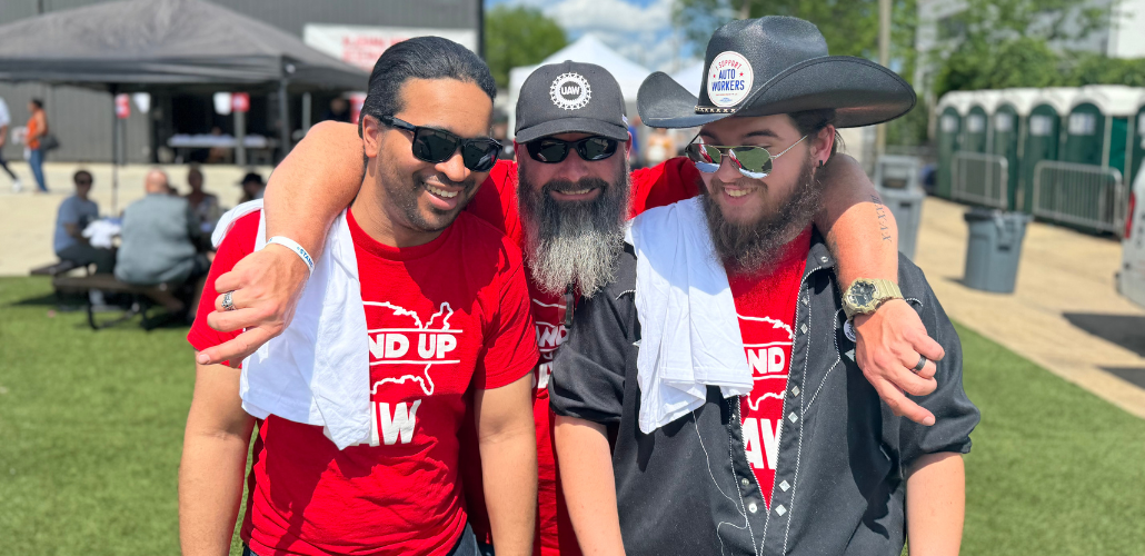 Three guys in red Stand-Up Strike (UAW) t-shirts, two bearded, stand outside on grass with arms around each other’s shoulders, smiling big.