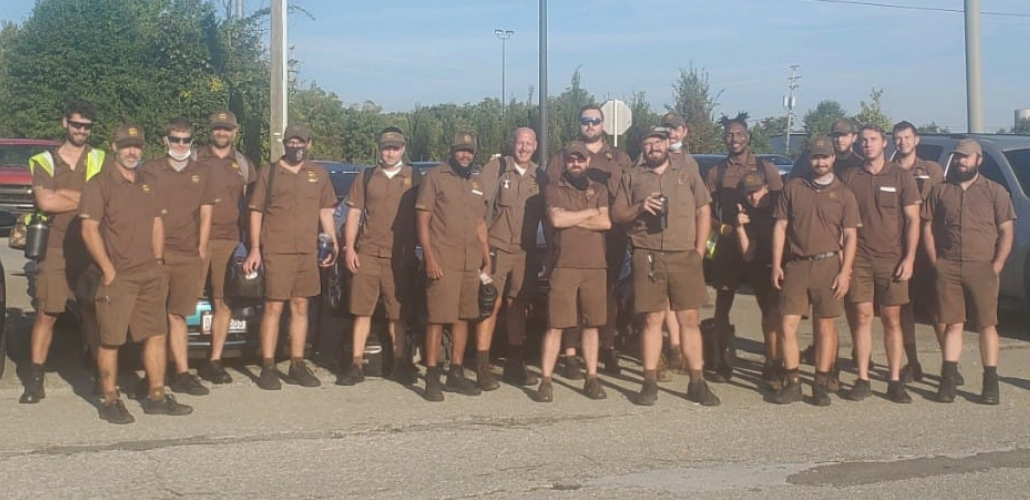 About 20 UPS workers in brown shorts uniforms pose for the camera together in a sunny parking lot. Many are smiling.