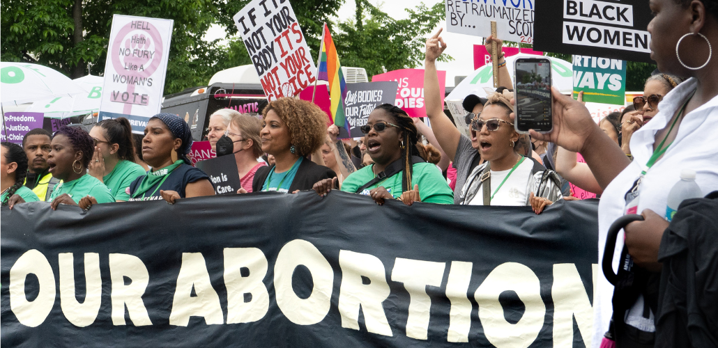 Women march behind a giant banner. The part we can see reads "OUR ABORTION." Other signs visible say "If it's not your body, it's not your choice" and "Our bodies, our future."