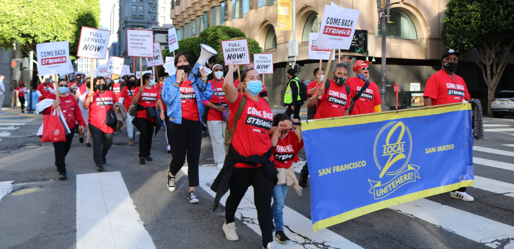 Workers march in downtown streets behind a big blue banner that says "UNITE HERE Local 2." They are wearing red T-shirts that say "Come back stronger" and some carry picket signs saying "Full service now!" or "Hotel rooms should be cleaned every day."  Most wear masks, one holds a bullhorn, and there is a child near the front of the crowd.