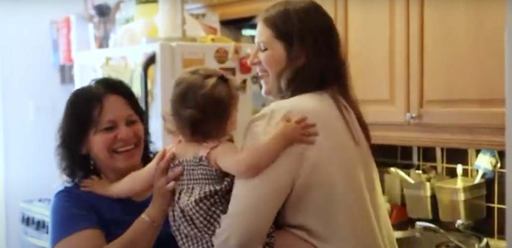 A white woman, right, hands a baby to a Latina woman, left. They are in an apartment kitchen. Both are smiling big. The baby's face is not visible.