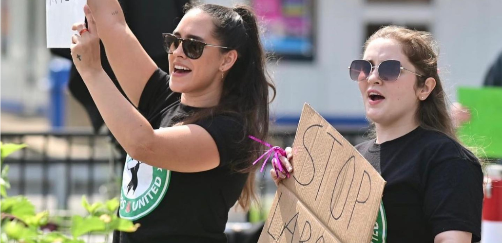 Two young women in Starbucks Workers United T-shirts stand outdoors, holding up cardboard signs, mouths open to yell or chant.
